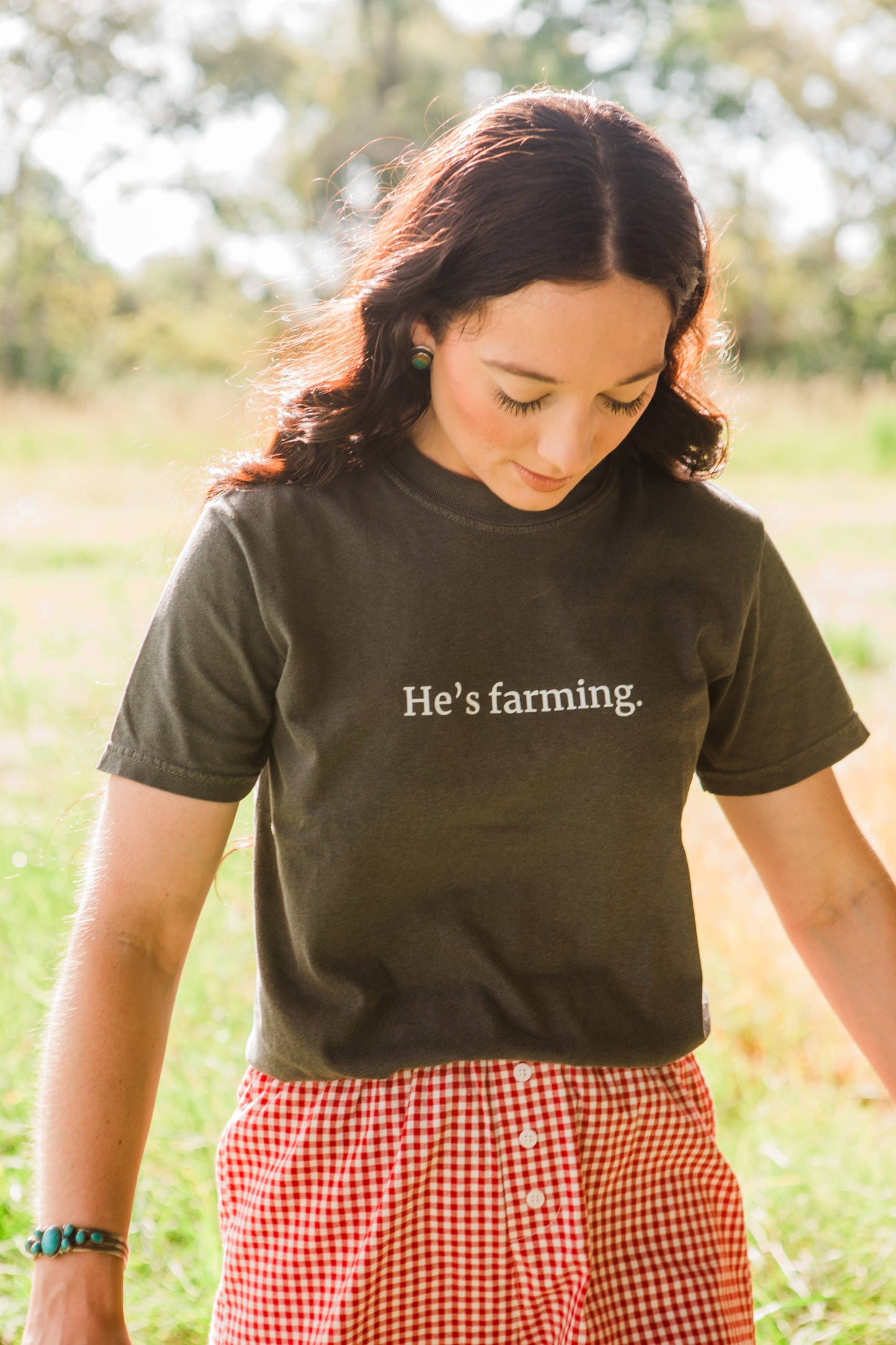 Woman wearing a grey t-shirt with 'He's farming.' text in a field