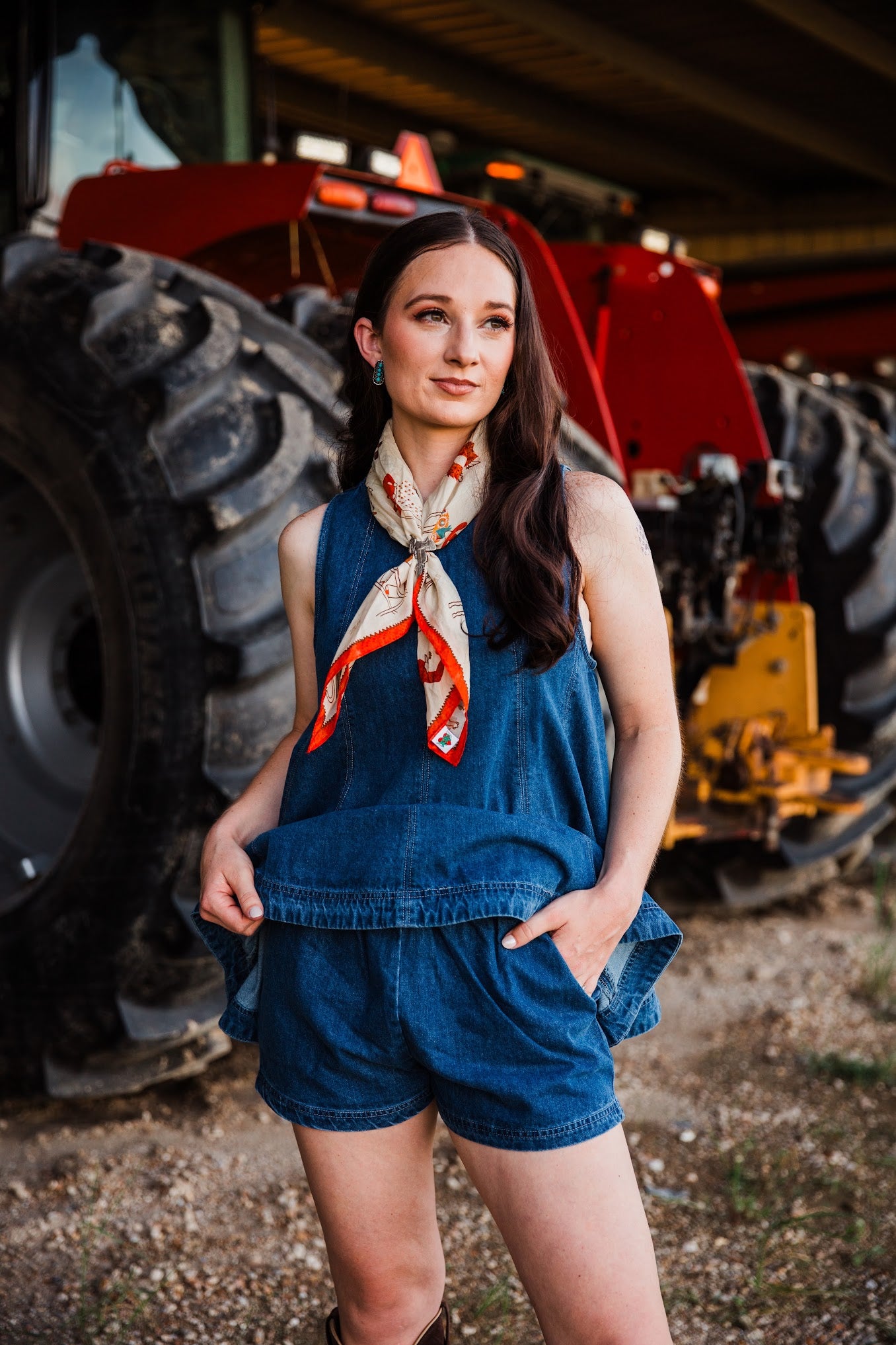 A woman wearing a blue denim sleeveless top and matching shorts, styled with a red and white patterned neck scarf, standing in front of a red tractor in a barn setting.