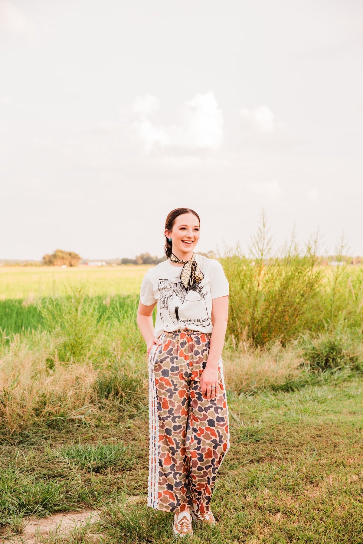 Smiling model in the Keepin’ It Western cream tee by XOXO Art & Co, paired with a floral neck scarf and patterned pants outdoors.
