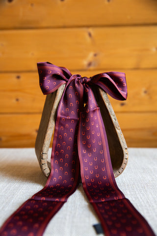 2 Fly Co My Lucky Twilly in Wine, a burgundy horseshoe-print scarf tied in a bow on a wooden stirrup with long draped ends.