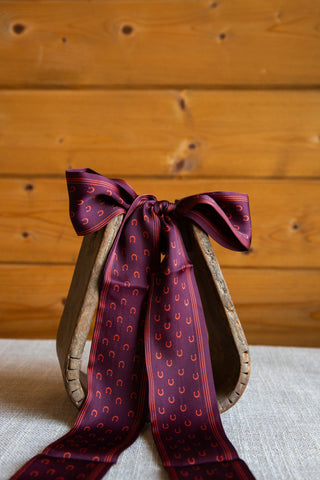 My Lucky Twilly in Wine by 2 Fly Co displayed on a wooden stirrup, with burgundy horseshoe-print tails beneath a knotted bow.