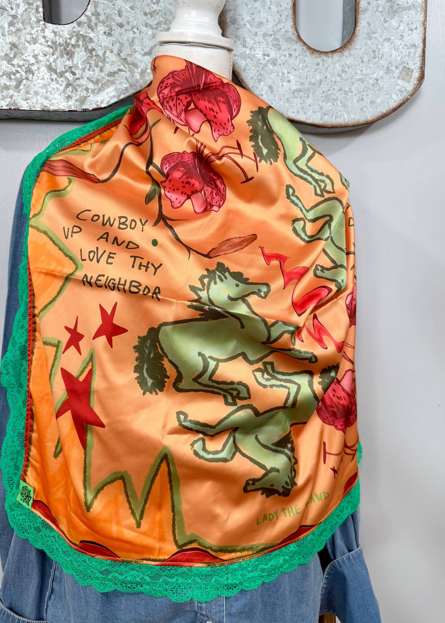 An orange silk scarf with a green and red western-themed print, featuring a horse, stars, and the text 'COWBOY UP AND LOVE thy NEIGHBOR', draped on a mannequin torso.