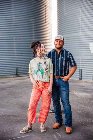 Sadie smiling toward Trevor while wearing the light sage Adios graphic tee with orange pants, a neck scarf and sneakers.
