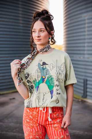 Sadie wearing the light sage Adios Tee with a colorful cowboy-on-bull graphic, styled with a printed neck scarf.