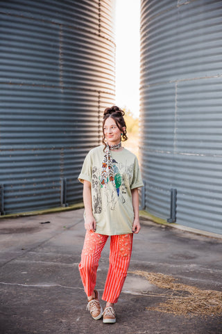 Full front view of the light sage Adios Tee showing its loose silhouette and Western graphic, paired with orange pants.
