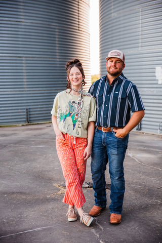 Sadie wearing the Adios Tee tucked into orange patterned pants, standing beside Trevor between metal grain bins.