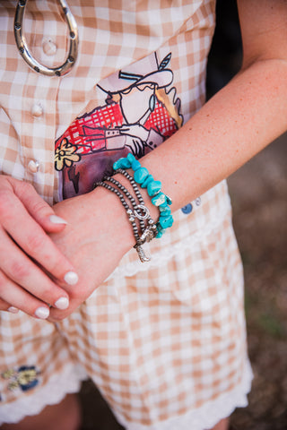 A close-up of a person's wrist wearing multiple bracelets, including turquoise beaded bracelets, silver-toned chain bracelets, and a small silver cross charm, paired with a peach-and-white gingham dress.