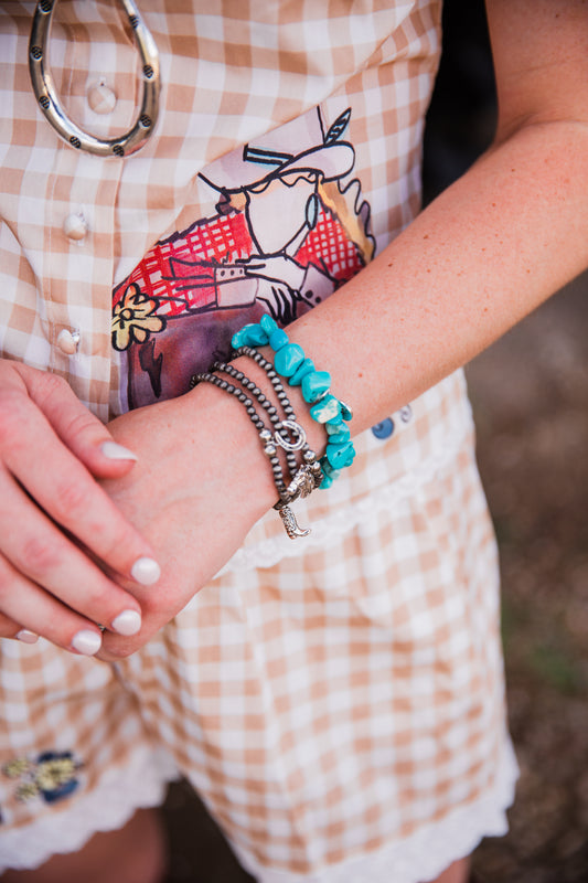 A close-up of a person's wrist wearing multiple bracelets, including turquoise beaded bracelets, silver-toned chain bracelets, and a small silver cross charm, paired with a peach-and-white gingham dress.