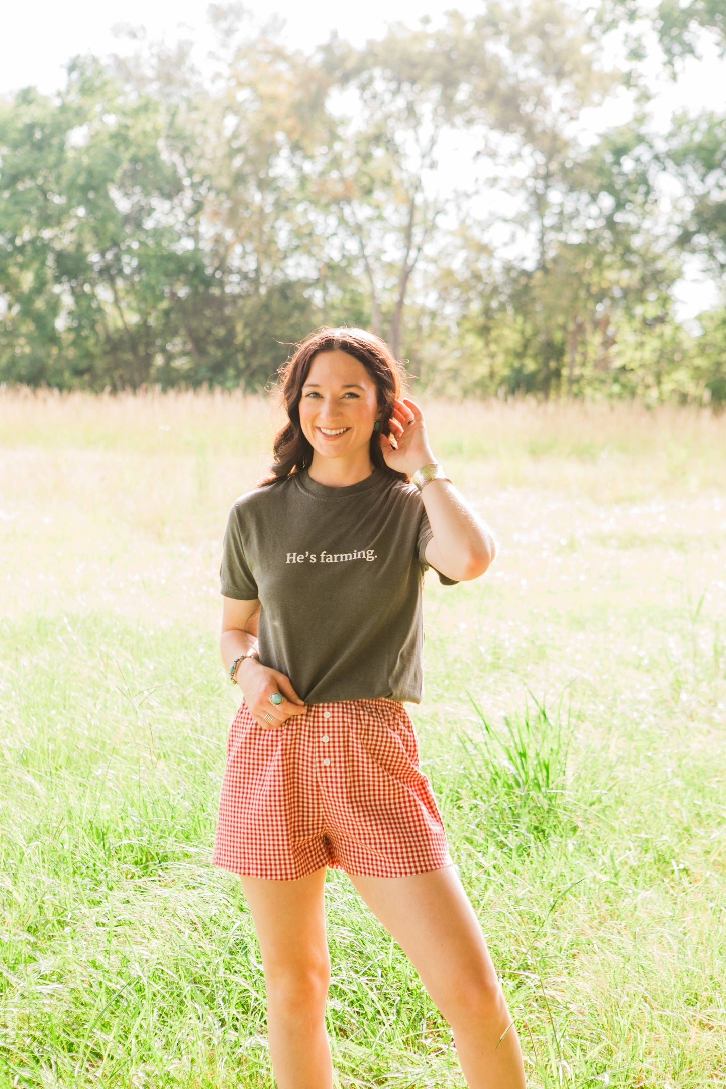 Woman standing in a field wearing a t-shirt that says 'he's farming' and red checkered shorts.