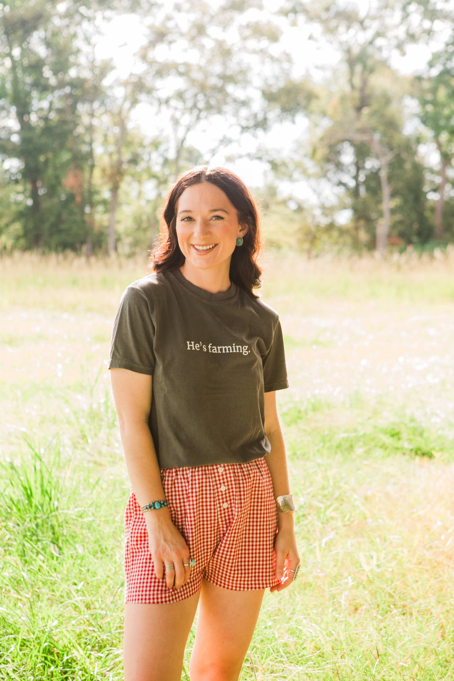Woman wearing a 'he's farming' t-shirt with text and red checkered shorts standing in a field.