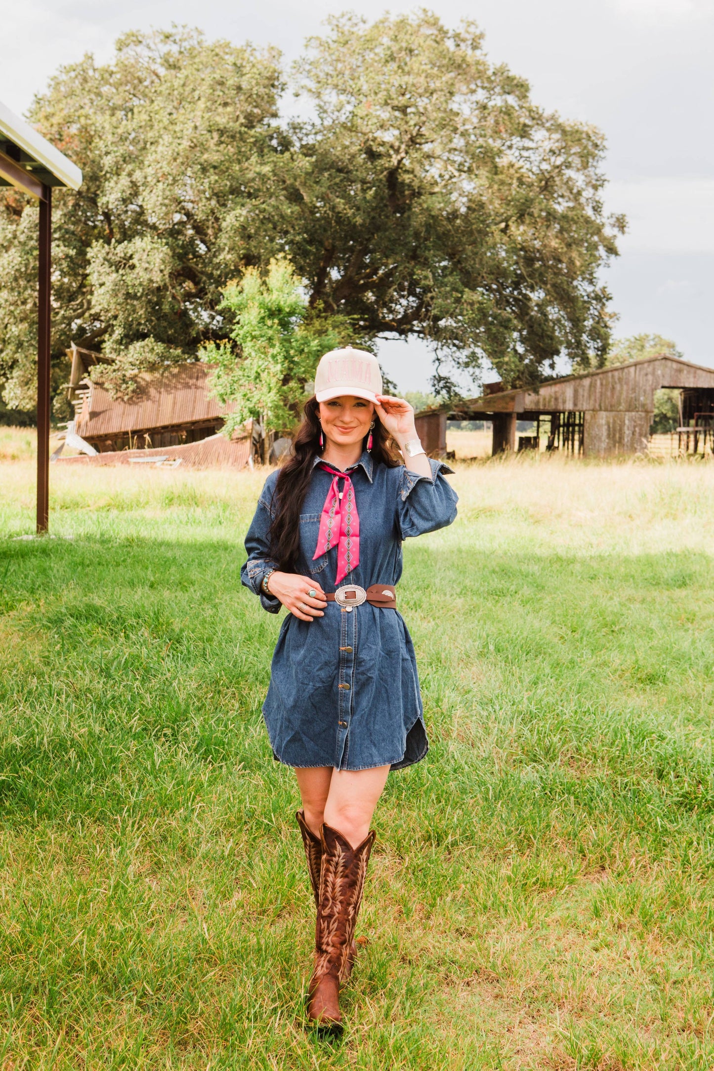 Woman in a denim dress and cowboy boots standing in a grassy field with trees and a wooden structure in the background.