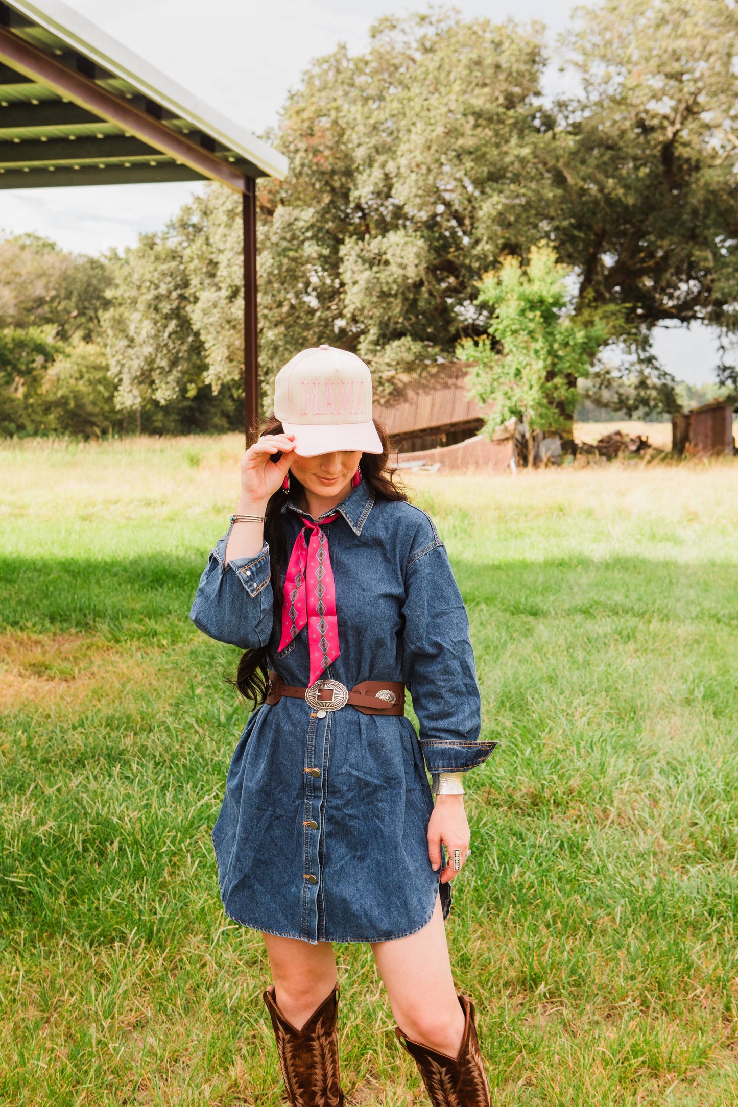 Woman in a denim dress and cowboy boots standing in a grassy field with trees in the background