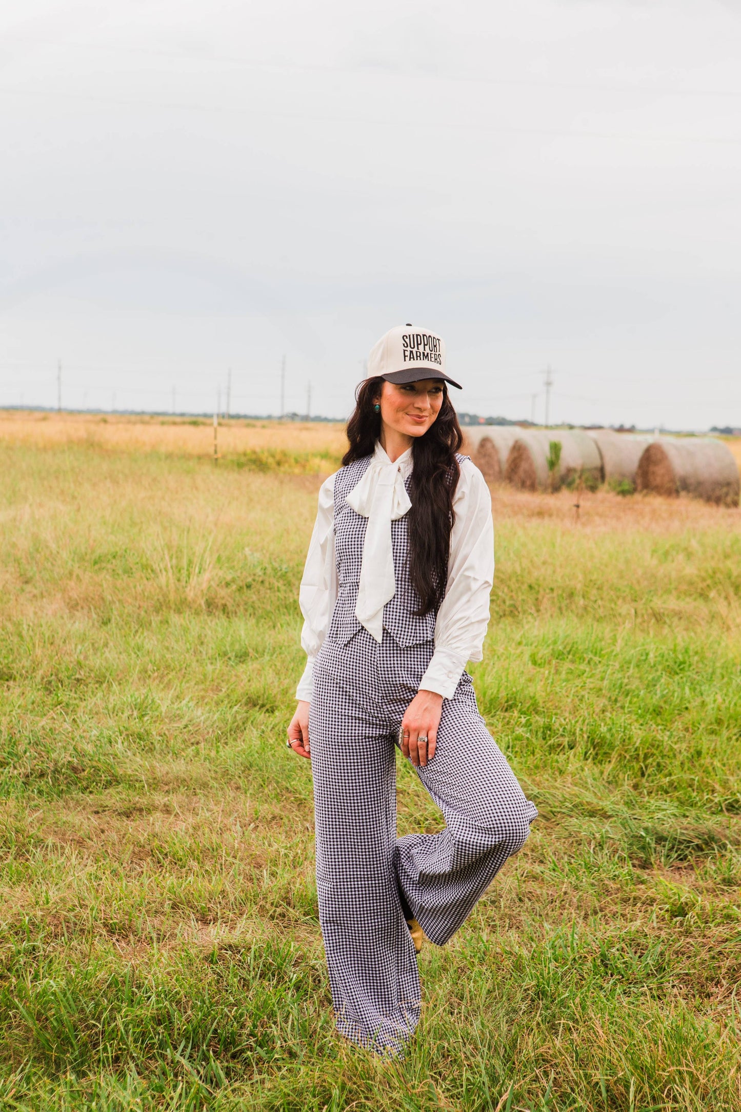 Person wearing a white shirt, black checkered pant set, and a cap in a field with hay bales.