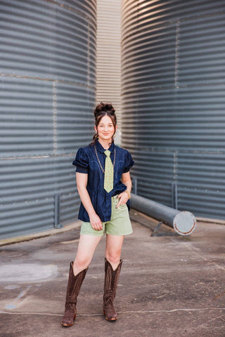 Full-length view of Sadie wearing the green Cowhand Tie with a dark denim top, green shorts and tall brown cowboy boots.