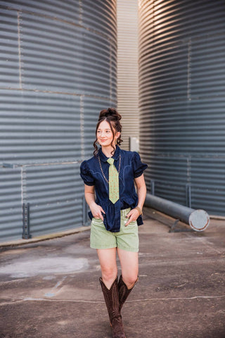 Sadie walking in the light green Cowhand Tie, styled with a dark denim puff-sleeve top, green shorts and brown cowboy boots.