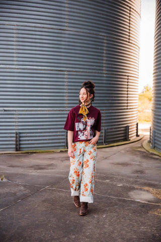 Sadie wearing the burgundy Dilly Dally Tee with light floral pants, brown boots and a gold scarf beside a metal grain bin.