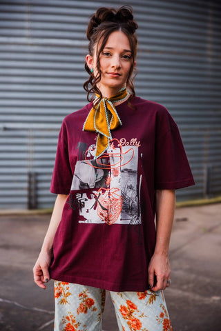 Sadie wearing the burgundy Dilly Dally Tee untucked, showing its Western collage graphic, styled with a gold neck scarf.