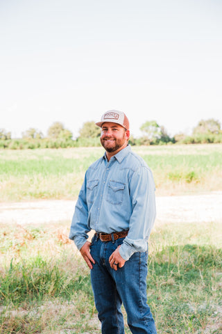 Model outdoors in a light wash denim pearl snap shirt paired with dark jeans, a tooled leather belt, and a trucker cap.