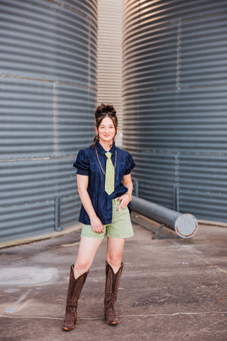 Full-length view of Sadie styling the High Roller denim top with a partial tuck, green shorts, a green tie and cowboy boots.