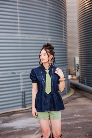 Sadie wearing the dark denim High Roller Top with puff sleeves and a gathered waist, styled with a green tie and shorts.