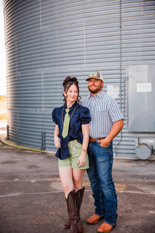 Sadie wearing the High Roller denim top with green shorts and tall brown cowboy boots, standing beside Trevor.