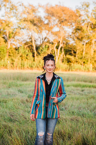 Front portrait of Sadie wearing the teal Jade Serape Blazer with striped lapels, a black Western blouse and ripped jeans.