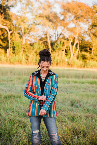 Sadie adjusting the sleeve of the teal striped Jade Serape Blazer, paired with a black blouse, red belt and blue jeans.