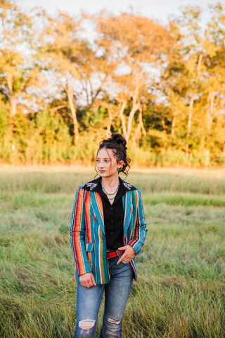 Sadie wearing the teal Jade Serape Blazer with multicolored stripes, styled over a black Western blouse and distressed jeans.