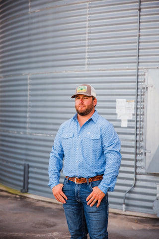 Trevor modeling the blue patterned Jesse long-sleeve pearl snap and jeans beside a metal grain bin.
