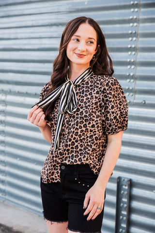 Model holding the striped scarf bow on the Lexington Leopard Bow Blouse, styled with black denim shorts.