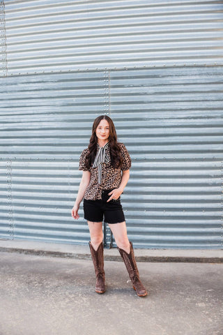 Model wearing the Lexington Leopard Bow Blouse with black denim shorts and tall brown Western boots.