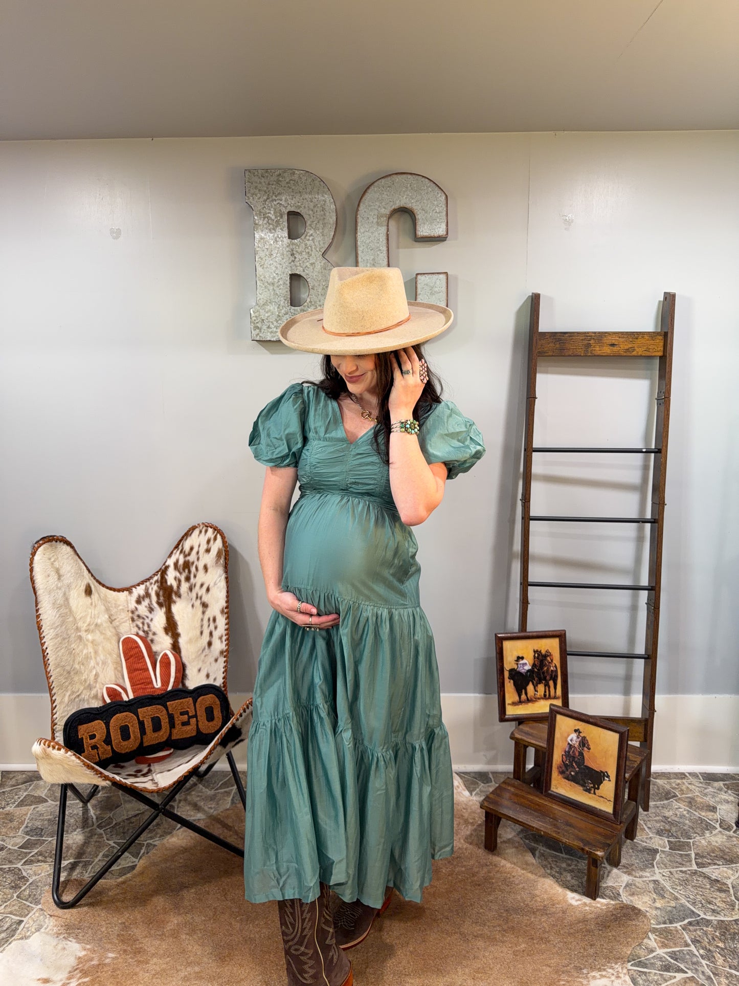 Woman in a teal dress and hat standing in a room with rustic decor, including a 'Rodeo' chair and wooden ladder.