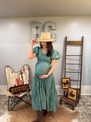 Woman in a teal dress holding a cowboy hat in a room with rustic decor.