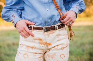 Close-up of Paisley Horse Pants showing cream fabric, horse print, front pockets and belt loops with a leather belt.