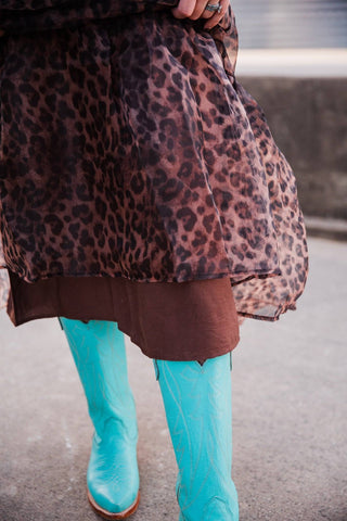 Close-up of the Sable Leopard Maxi Dress’s sheer leopard-print organza hem over a brown underlayer and turquoise boots.