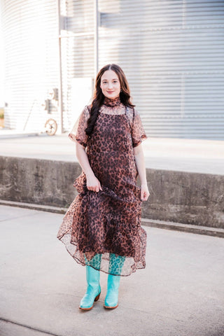  Model lifting the Sable Leopard Maxi Dress’s ruffled skirt slightly, styled with turquoise knee-high Western boots.