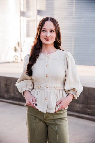 Sadie looking to the side in the cream Sutton Top, showing its button front, gold-tone waist accents and puff sleeves.