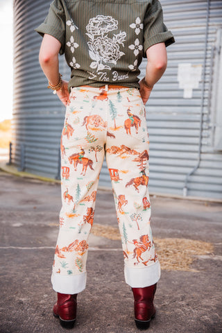Back view of the cream Cowboy Way Pants showing the Western scenic print and rolled cuffs above burgundy boots.