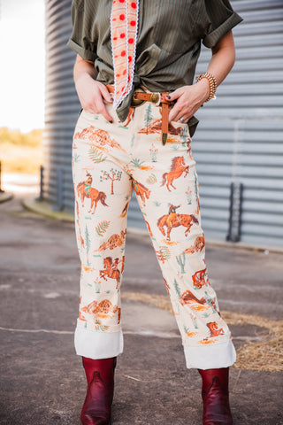Front view of the cream Cowboy Way Pants showing cowboy, horse and desert prints, styled with rolled cuffs and burgundy boots.