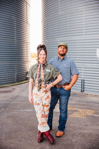 Sadie modeling the cream Cowboy Way Pants with an olive shirt and burgundy boots, standing beside Trevor between grain bins.