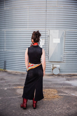 Back view of the fitted black denim Walk the Line Dress showing its sleeveless shape and back slit, paired with red boots.