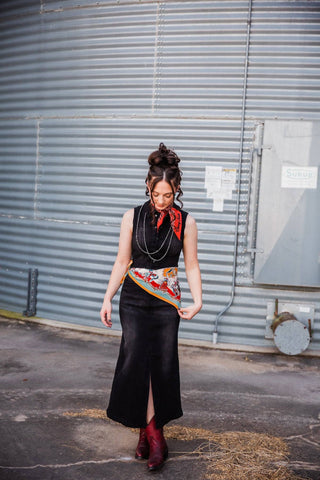 Black denim Walk the Line Dress showing its front skirt slit, styled with a printed waist scarf and red Western boots.