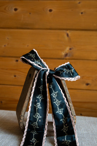 Devotions silk twilly by Allie Falcon tied in a bow, featuring a navy background, light ornamental print and pale crochet trim.