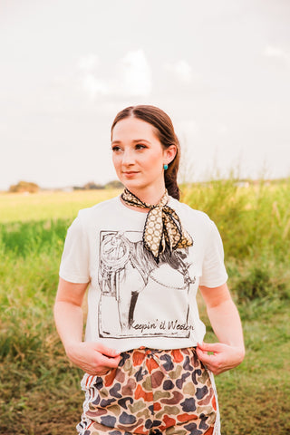 Josephine Twilly by Allie Falcon worn as a neck bow with a cream horse graphic tee and multicolor patterned pants outdoors.
