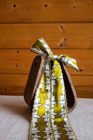 Mod Maud silk twilly by Allie Falcon tied in a bow, with a yellow and white cowboy-hat profile print and crochet trim.