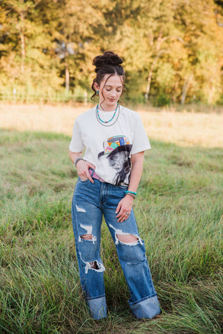 Front view of the white Reverie Tee by Allie Falcon tucked into cuffed jeans, featuring a cowboy portrait and colorful hat.