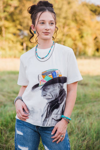 Allie Falcon Reverie Tee in white, front view of a cowboy portrait with a colorful patterned hat, styled with blue jeans.