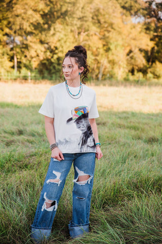 Reverie Tee by Allie Falcon styled outdoors with cuffed jeans, showing the white short-sleeve shirt and western cowboy artwork.