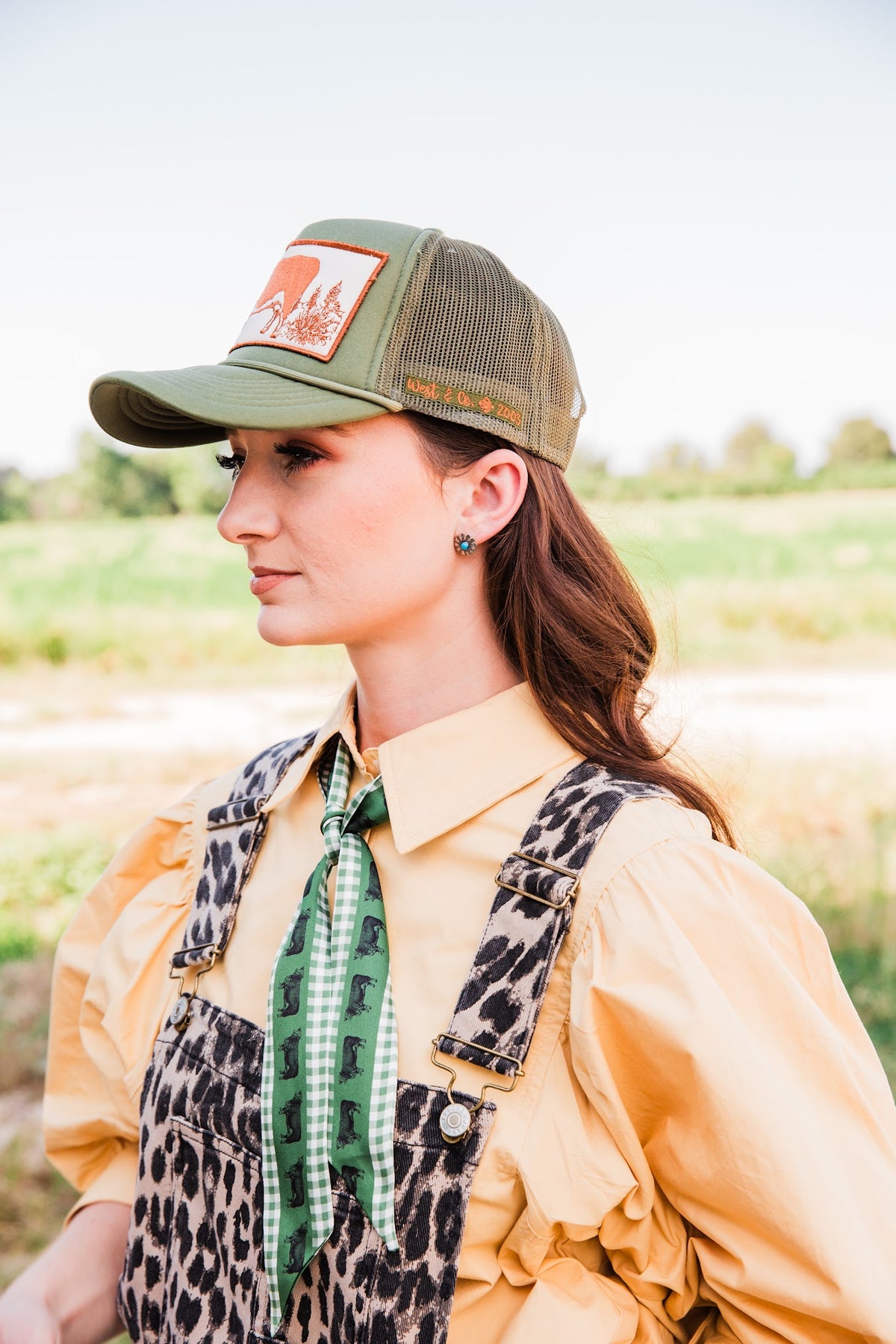 Model wearing a turquoise concho stud earring with a green trucker hat, tan blouse, and leopard overalls in a side view.