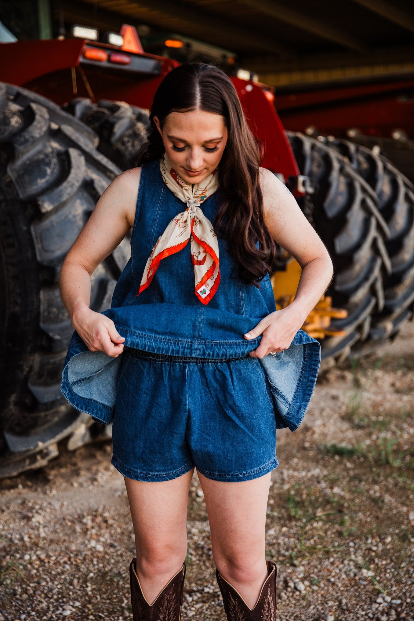 A woman wearing a denim sleeveless top and matching denim shorts stands in front of large farm tires, holding up the shorts to show the lining.